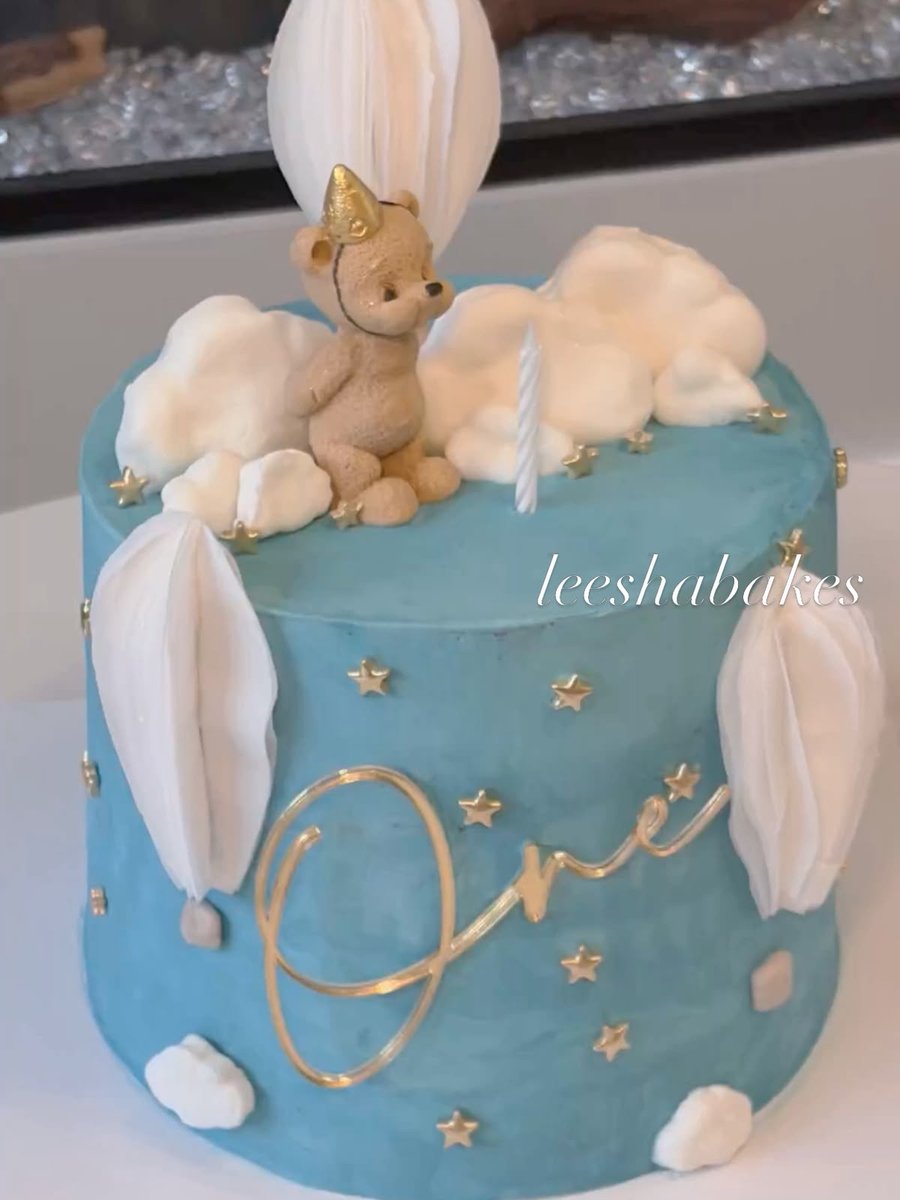 Blue first-birthday cake with a teddy bear topper, fluffy clouds, gold stars and gold 'One' lettering
